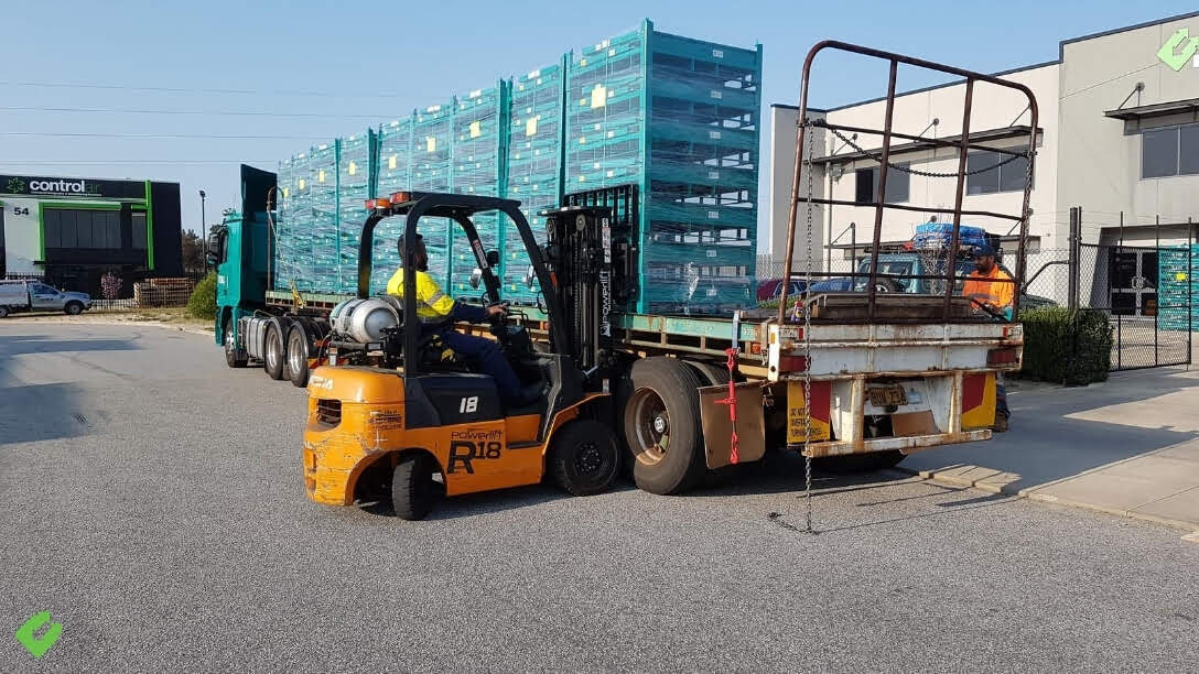 Backsafe forklift operator unloading stacked custom pallet cages from a flatbed truck outside the company’s warehouse facility.